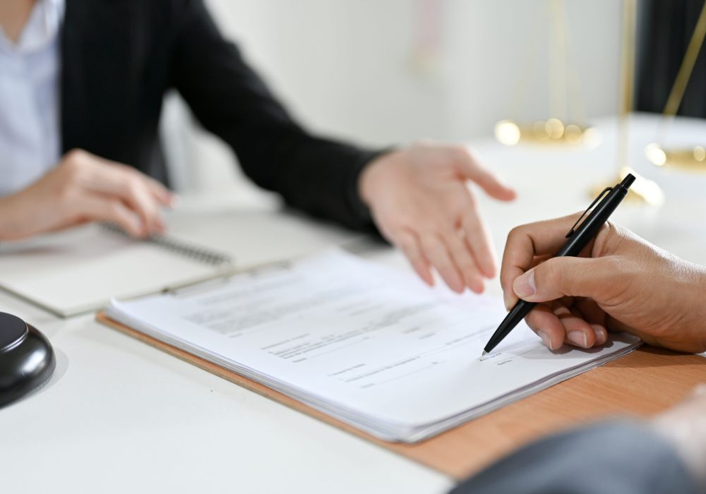Professional signing legal document in office setting, discussing terms with advisor over a desk, scales of justice blurred.