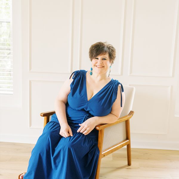 Kelly Brooks in blue dress sitting in a wooden chair, bright room background, relaxed and cheerful.