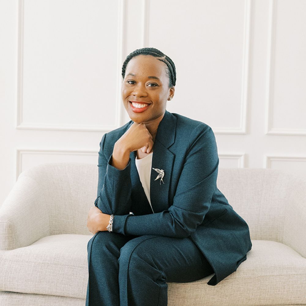 Professional woman in blue suit smiling while seated on a white sofa in bright, elegant office space.