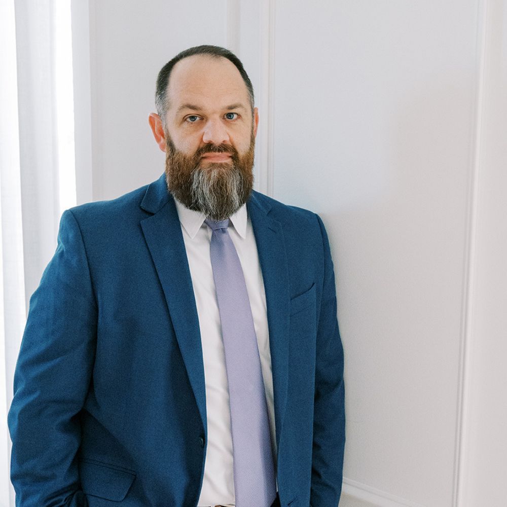 Thomas Clark in a blue suit with a beard and lavender tie, standing against a white wall, looking confident.