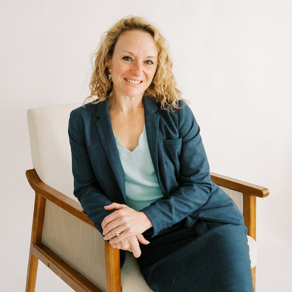 Brittany Pinkham smiling in a blue suit sitting on a chair, professional studio portrait.