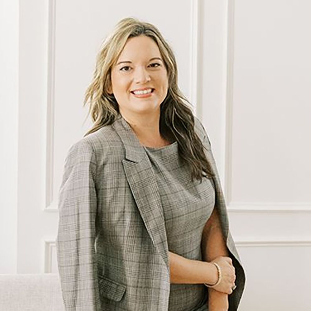 Ellie Bragg in a gray suit standing against a white wall background, professional portrait photography.