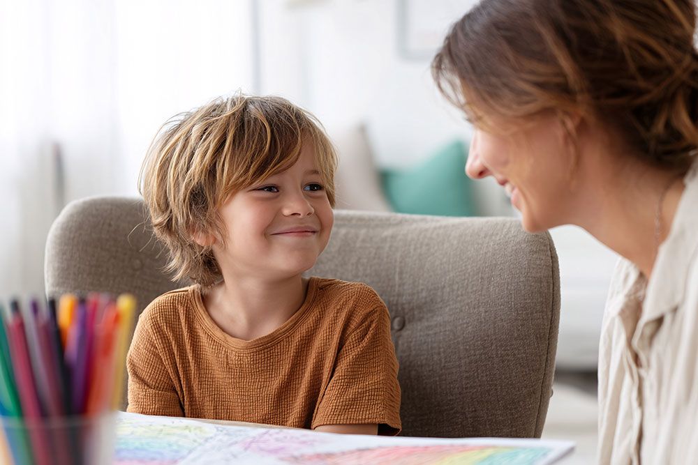 Smiling child in a brown shirt looks at a woman, with colorful pencils on the table in a cozy room.