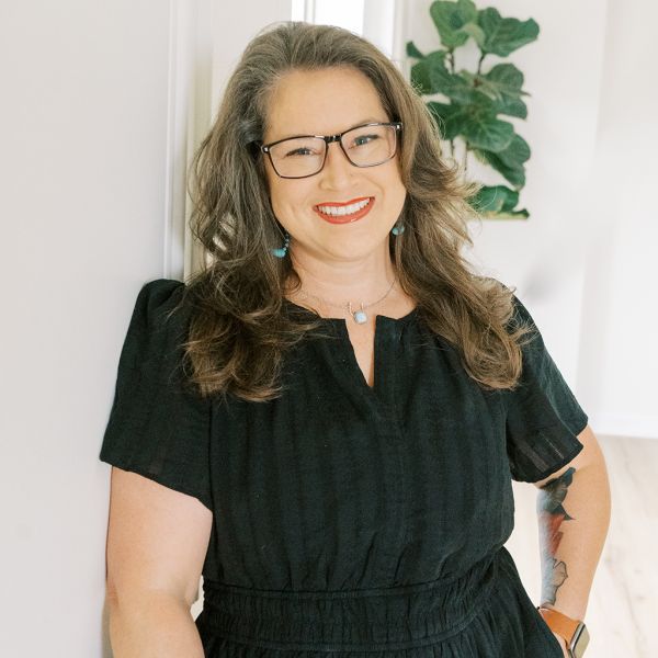 Jessica Hinson in black dress with glasses stands by white wall, plant in background.