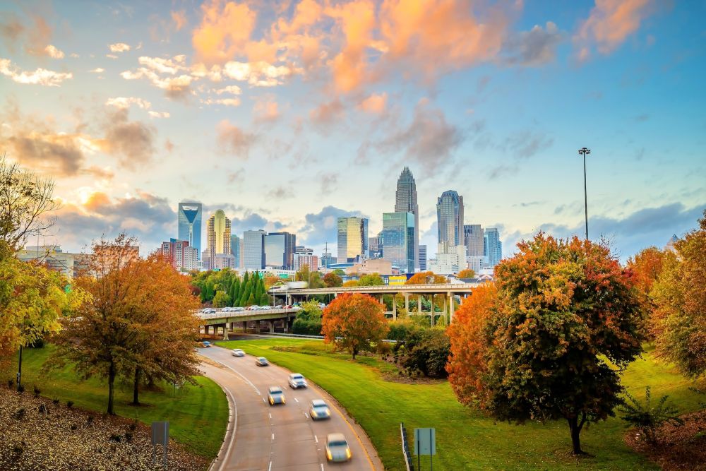 Autumn cityscape of Charlotte skyline with vibrant trees and a highway under a colorful sky.