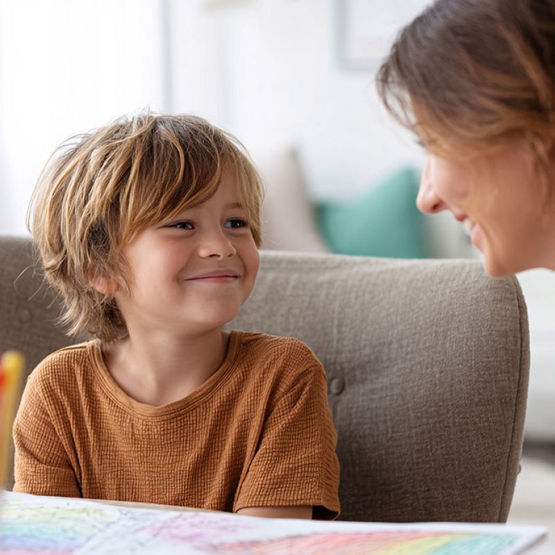 Smiling child in a brown shirt looks at a woman, with colorful pencils on the table in a cozy room.