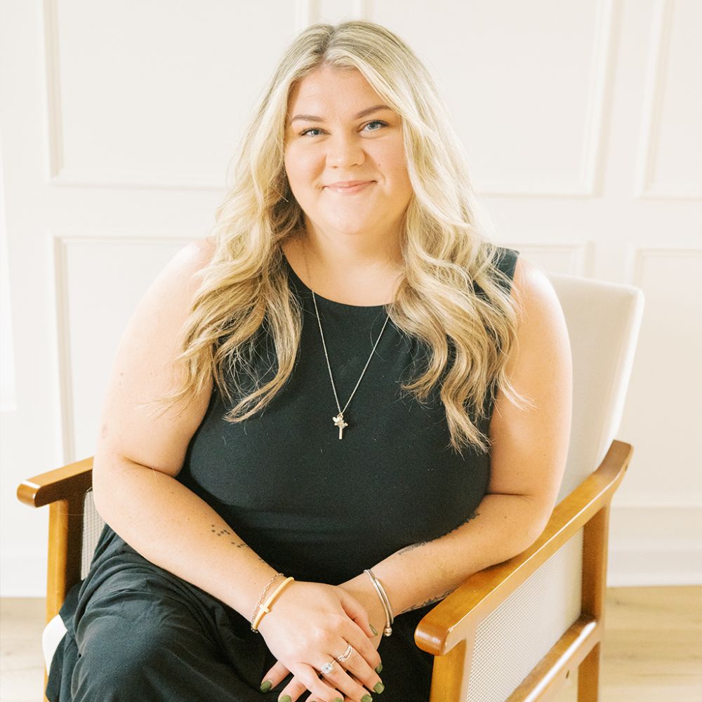 Caroline Pierce with long blonde hair in a black dress seated on a wooden chair in a bright room.
