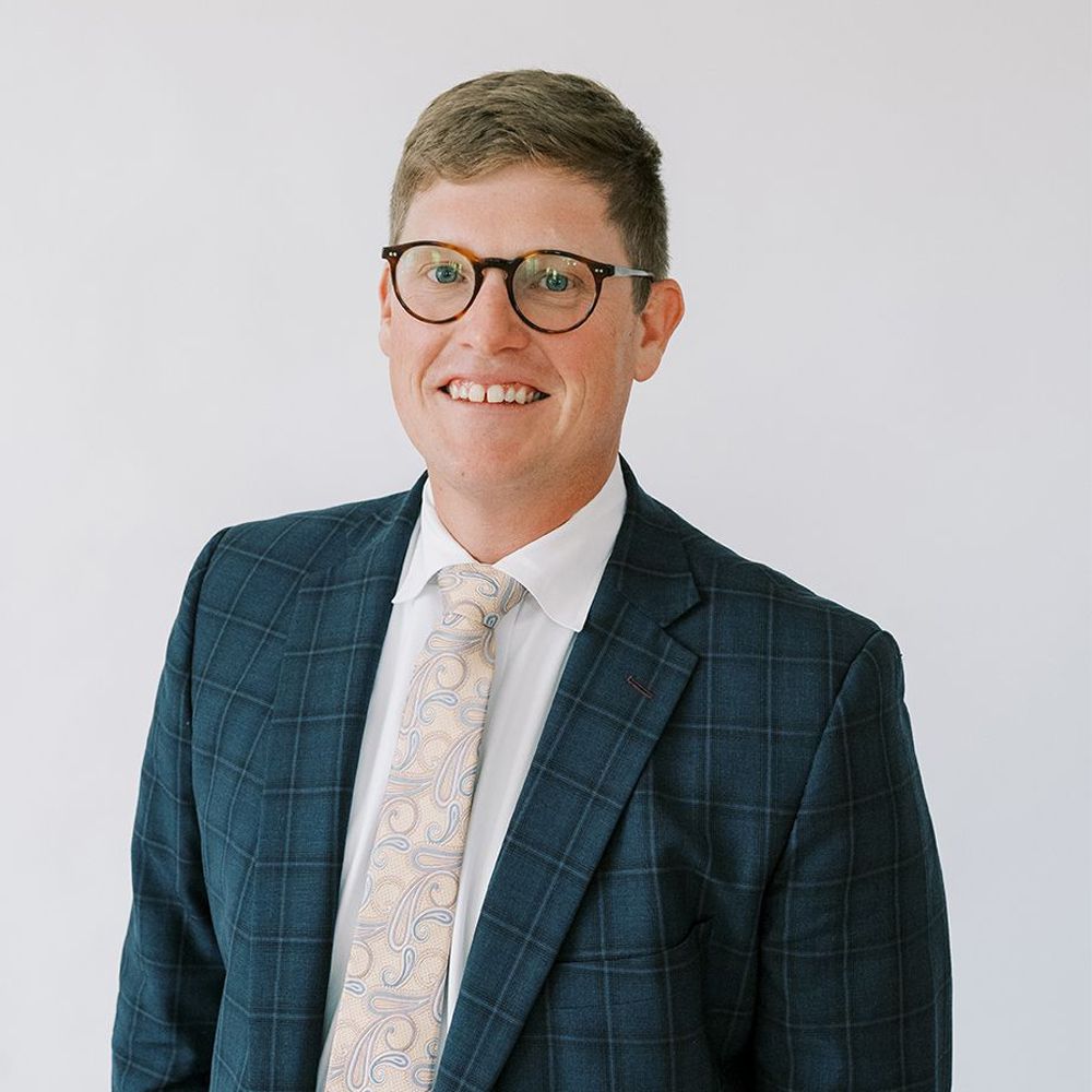 Tee Leitner in suit with glasses smiling against a plain background. Business portrait, professional attire.