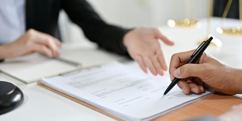 Professional signing legal document in office setting, discussing terms with advisor over a desk, scales of justice blurred.
