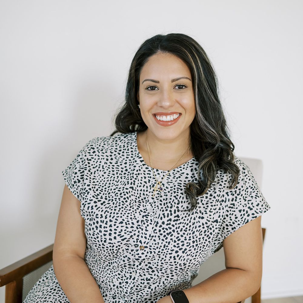 Cristal Verano in a patterned dress sitting on a chair, wearing a smartwatch and jewelry.