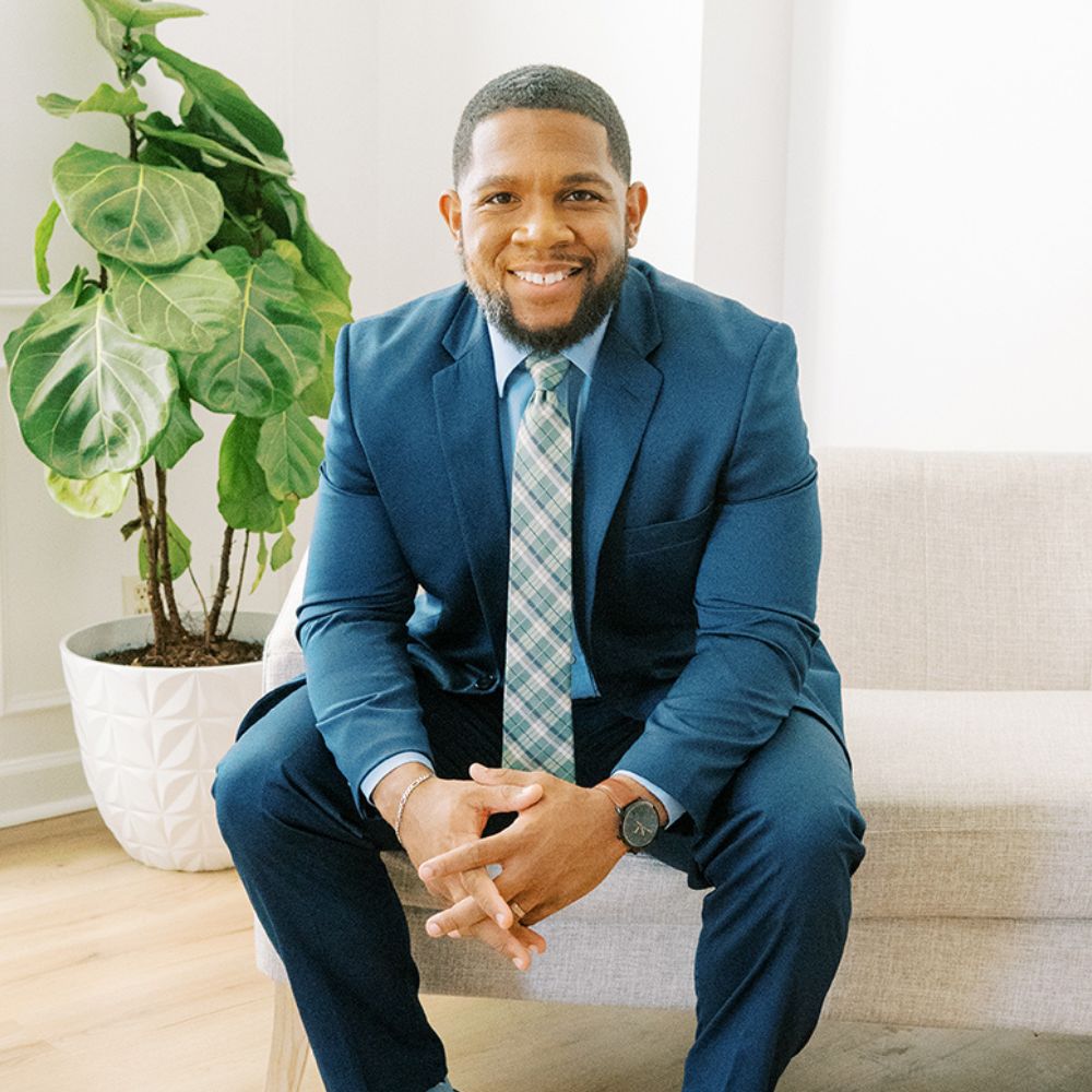 Bryan Sumner in blue suit sitting on sofa, smiling, next to green plant in bright room.
