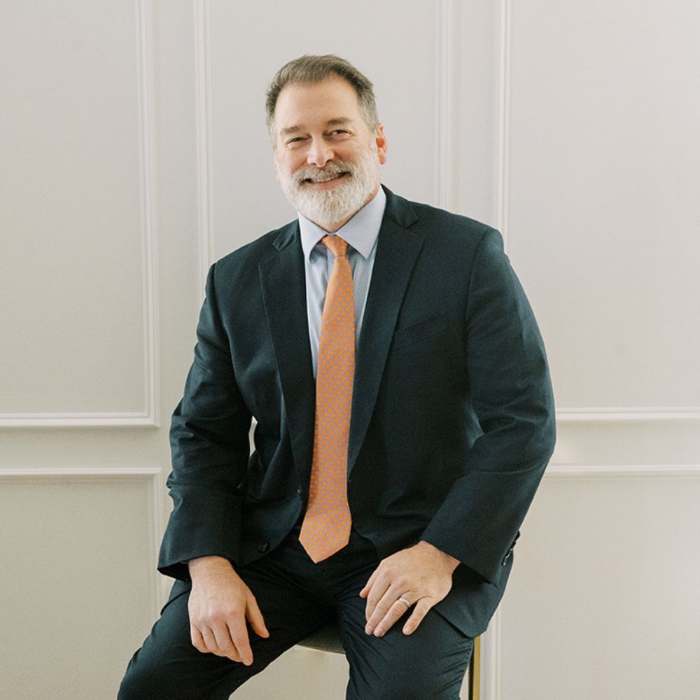 Ken Harris in suit with orange tie sits on a chair against a white paneled wall, smiling confidently.